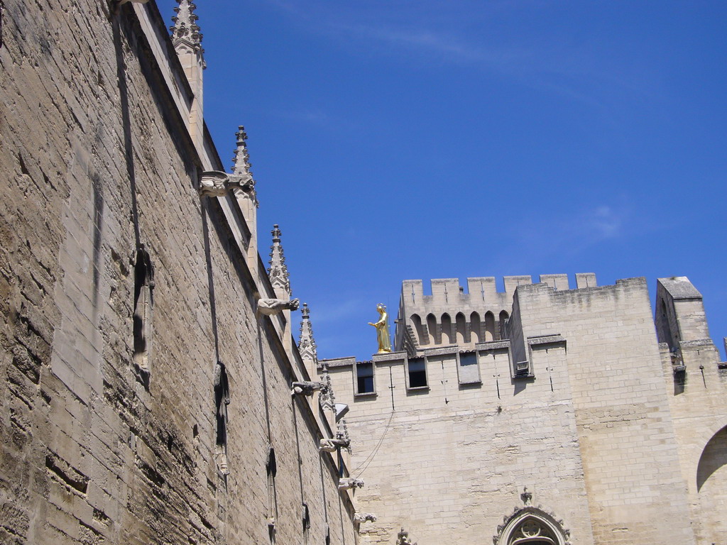 Walls at the Cour d`Honneur courtyard and the Tour de la Campane tower of the Palais des Papes palace, and the Gilded statue of the Virgin Mary at the top of the Avignon Cathedral