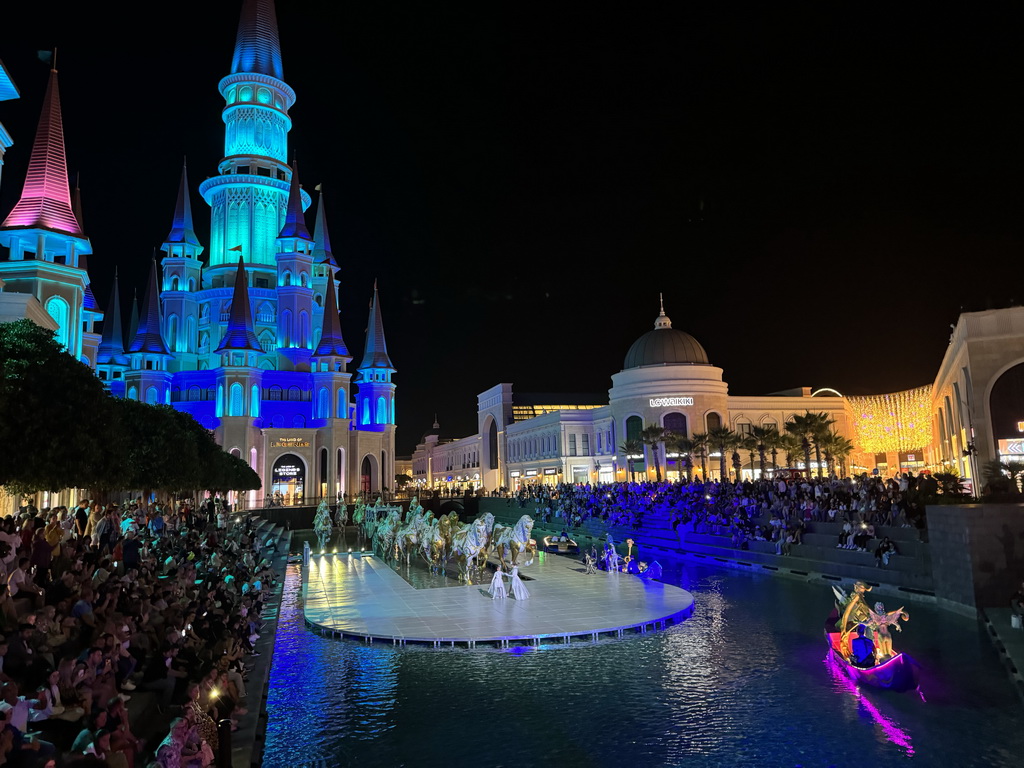 Boats, actors and horse statues in front of the Chateau at the Shopping Avenue area of the Land of Legends theme park, during the Musical Boat Parade, by night