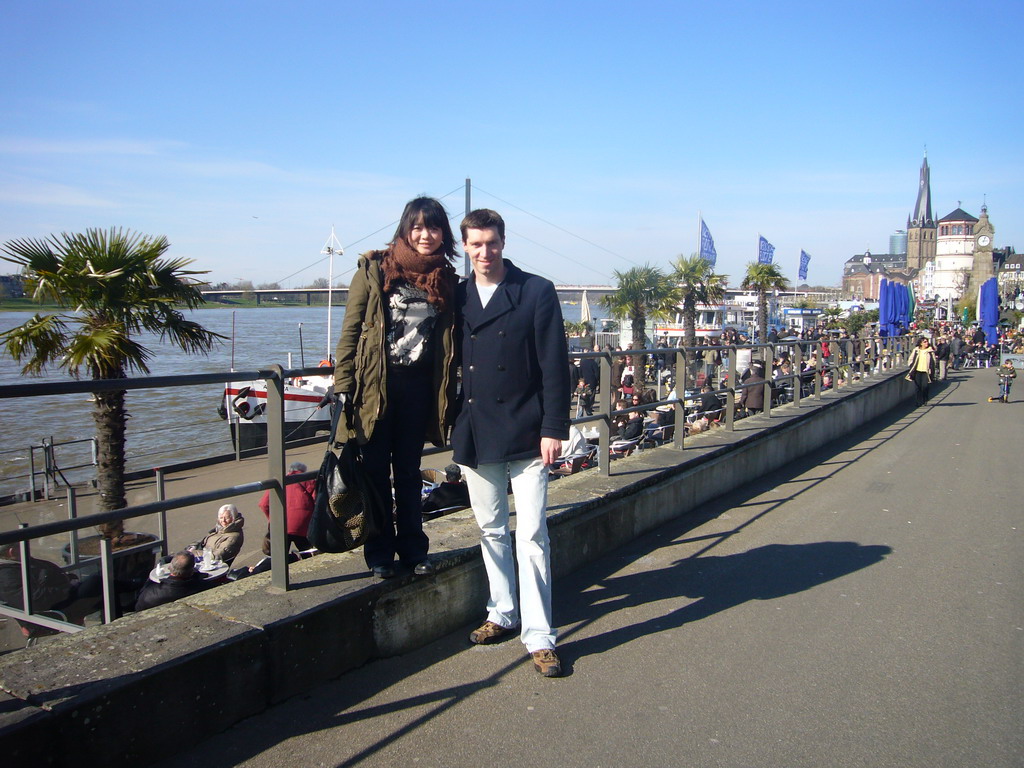 Tim and Miaomiao at the Rhine shore, with the Oberkasseler Brücke bridge, the Pegeluhr clock, the Old Castle Tower and the Lambertuskirche church