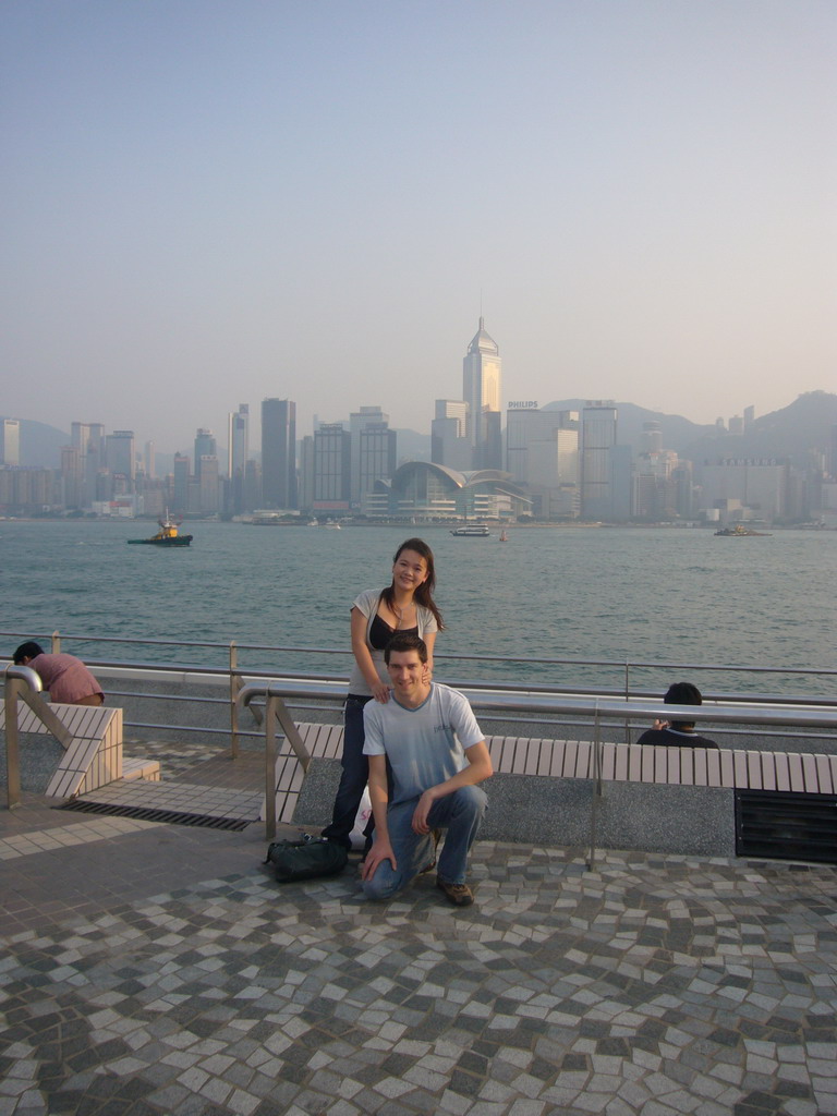 Tim and Miaomiao at the Kowloon Public Pier, with a view on boats in Victoria Harbour and the skyline of Hong Kong with the Central Plaza building and the Hong Kong Convention and Exhibition Centre