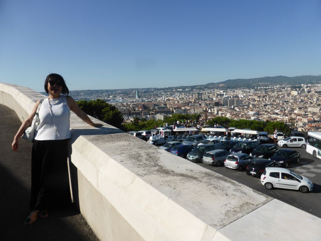 Miaomiao at the road to the Notre-Dame de la Garde basilica, with a view on the square in front with a tourist train and wooden cross, and the city center