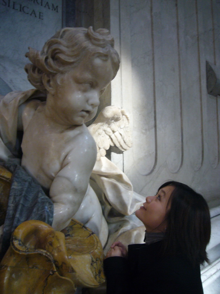 Miaomiao and the statue of a Cupido offering holy water, at the foot of the Monument of Pope Benedict XIII, inside St. Peter`s Basilica