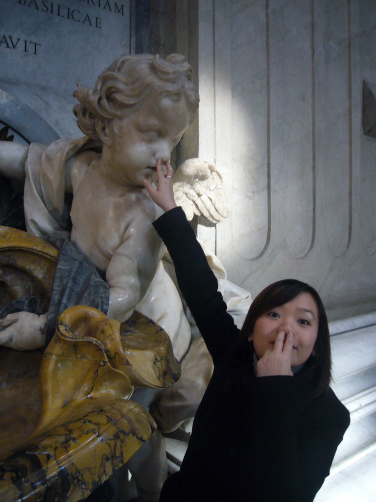 Miaomiao and the statue of a Cupido offering holy water, at the foot of the Monument of Pope Benedict XIII, inside St. Peter`s Basilica