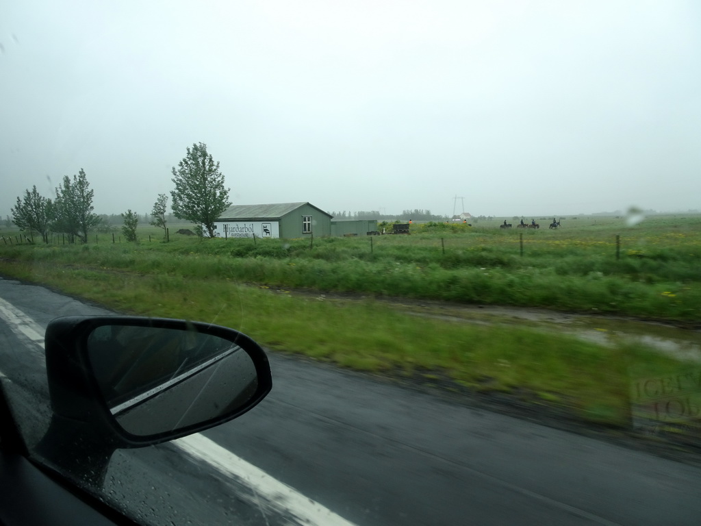 The Hjarðarból Guesthouse and people riding horses at Hjarðarból, viewed from the rental car on the Suðurlandsvegur road