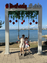 Miaomiao and Max on a swing at the square at Nar Beach 1, with a view on boats in the Gulf of Antalya and beaches and hotels at the northwest side of town